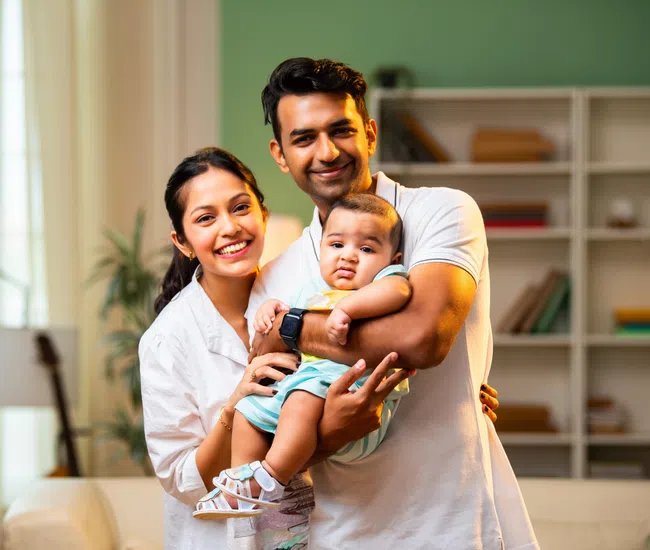Happy Indian family smiling at camera holding their baby in a bright cozy living room.
