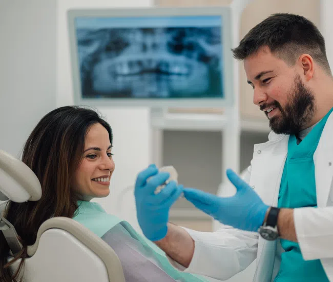 Dentist explaining a dental model to a smiling female patient in a dental clinic with X-ray on screen.