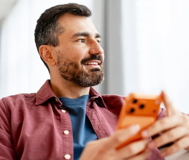 Smiling man with beard holding orange smartphone looking out bright window indoors relaxed and happy
