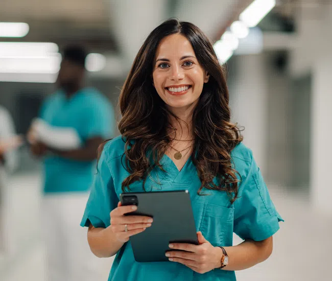 Smiling female nurse in blue scrubs holding a tablet with two medical professionals blurred in background