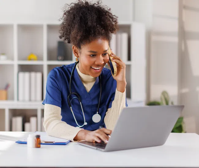 Smiling female nurse in blue scrubs talking on phone while working on laptop in bright office.
