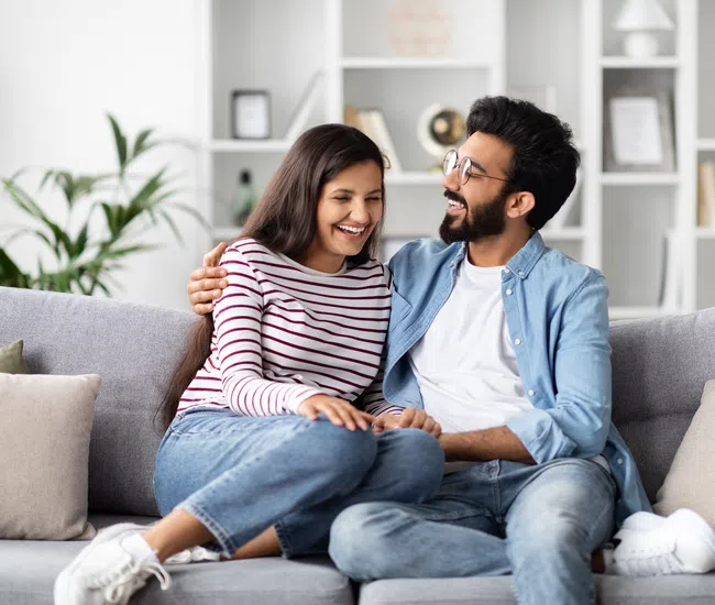 Happy young couple sitting on a sofa, smiling and embracing in a cozy modern living room with cushions and shelves.