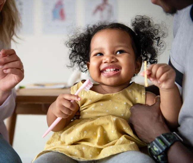 Smiling toddler in yellow dress holding toothbrushes with adults showing dental care in background