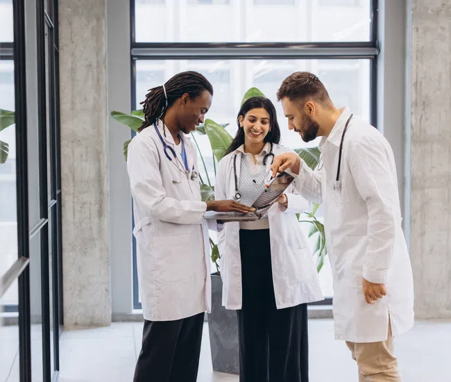 Three doctors in white coats discussing medical charts in a modern hospital hallway.