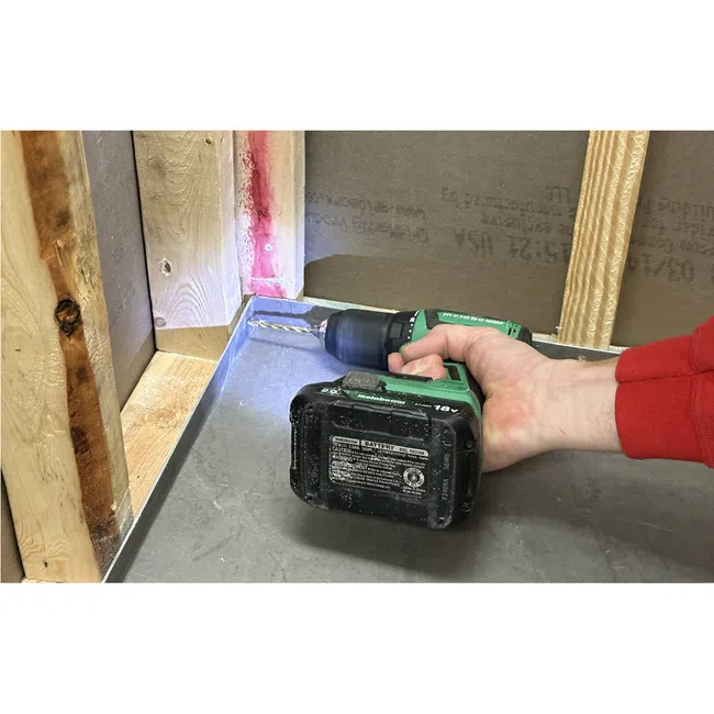 Hand holding green cordless drill with LED light, drilling into metal surface near wooden studs in construction.