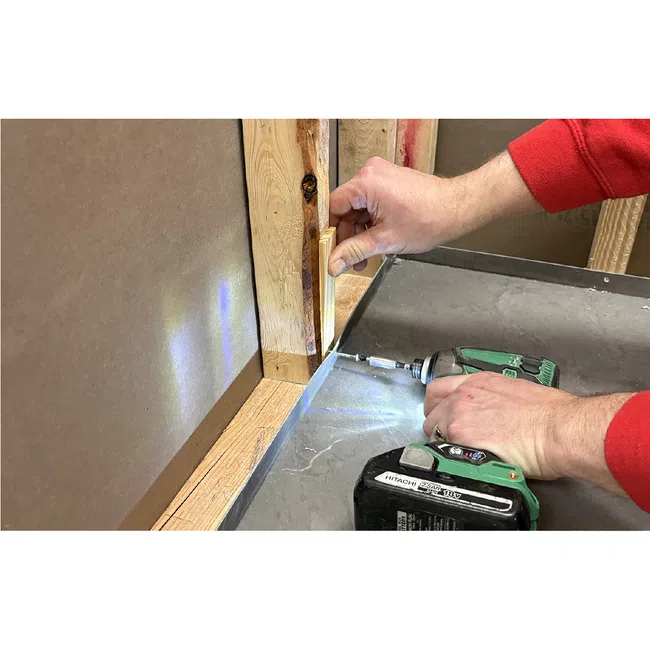 Person using a cordless drill to fasten wooden trim on a wall corner with drywall and studs visible.