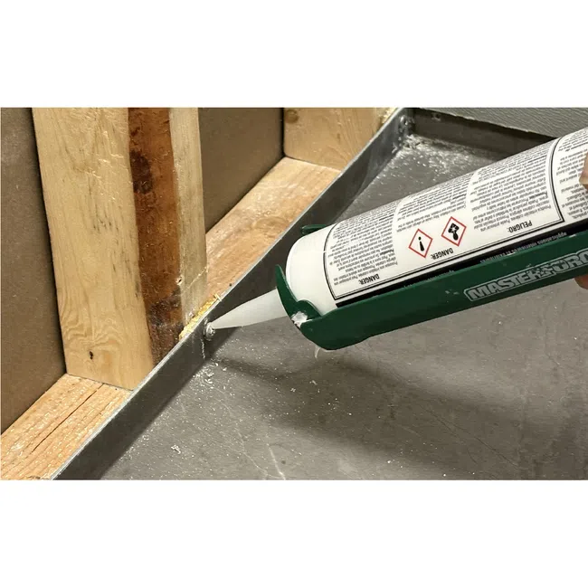 Applying white sealant with a caulking gun along the base of a wooden frame on a concrete floor.
