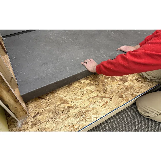 Person in red lifting or placing a large gray tile over oriented strand board flooring during installation.