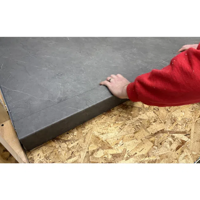 Person installing a large gray stone countertop on a wooden particleboard base wearing a red sleeve garment.