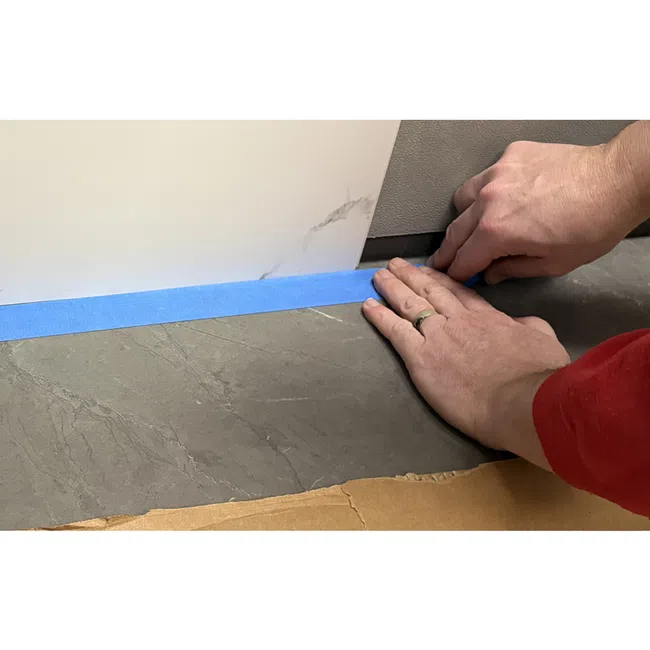Hands applying blue painter's tape along edges of a wall and gray tile floor for protection.