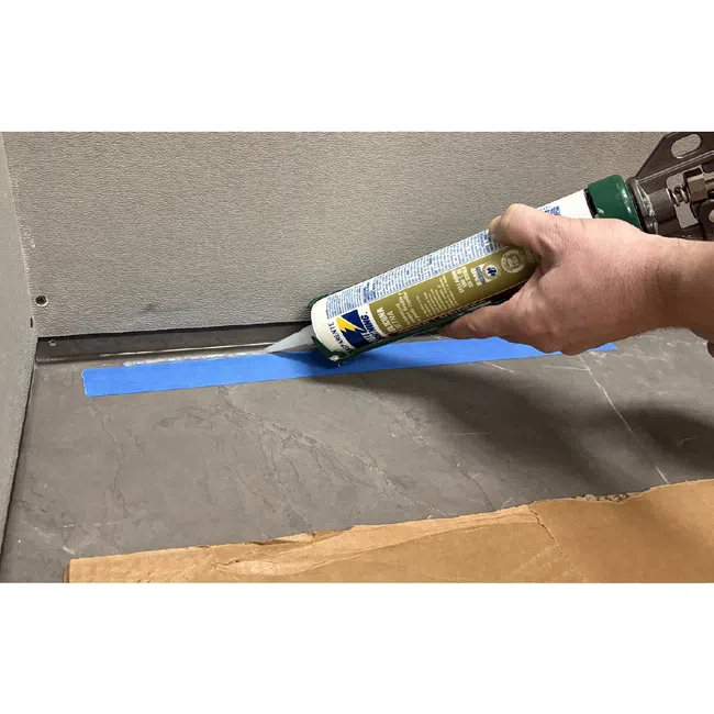 Hand applying white sealant along blue painter's tape on gray tile floor against wall corner