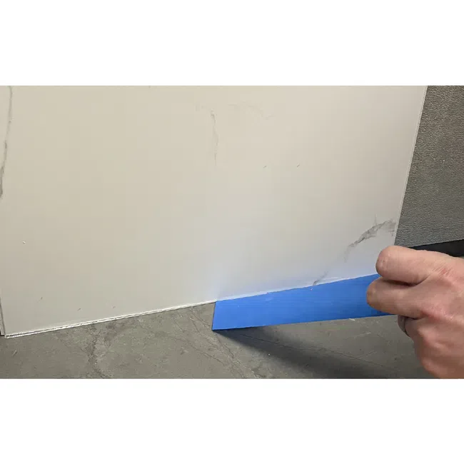 Hand holding blue scraper removing residue from the edge of a white surface on a grey floor.