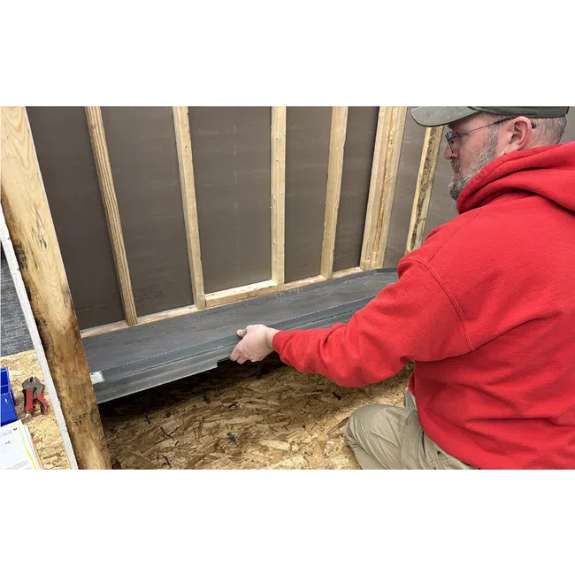 Man in red hoodie installing black material on wooden frame inside construction area with plywood floor.
