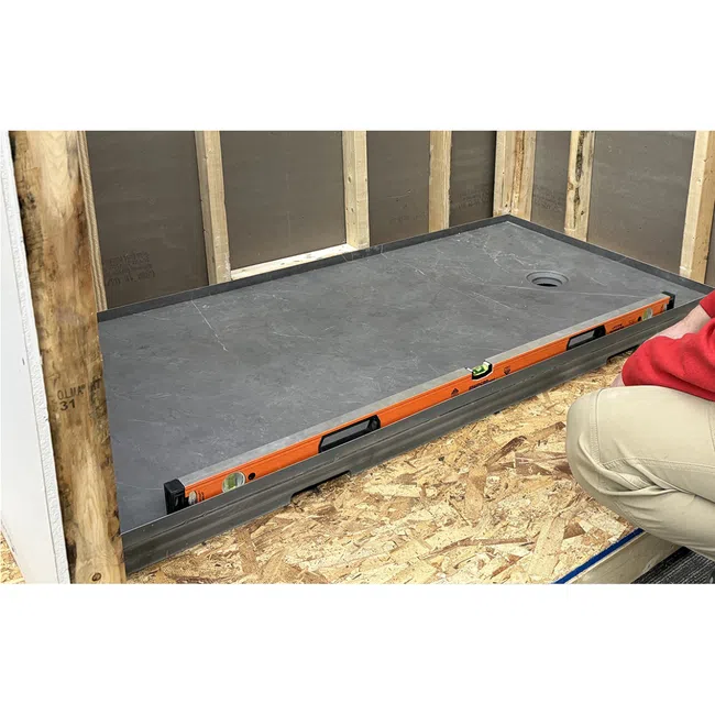 Worker installing a gray stone shower base with drain and level tool in unfinished wooden frame bathroom.