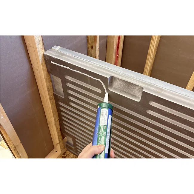 Hand applying adhesive sealant with caulking gun to the back of a shower base panel in wooden frame construction.