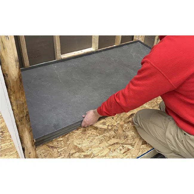 Person in red sweater installing large gray floor tiles on wooden subfloor in room under construction.