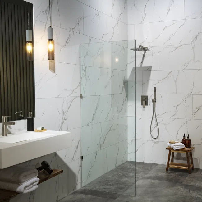 Modern bathroom with marble tiles, walk-in glass shower, wall-mounted sink, and wooden stool with towels.