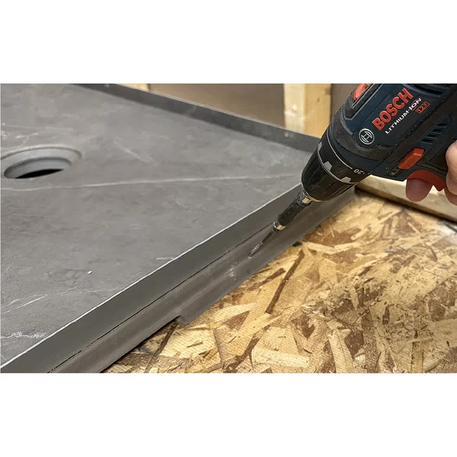 Handheld Bosch drill screwing into edge of gray stone countertop on plywood surface.