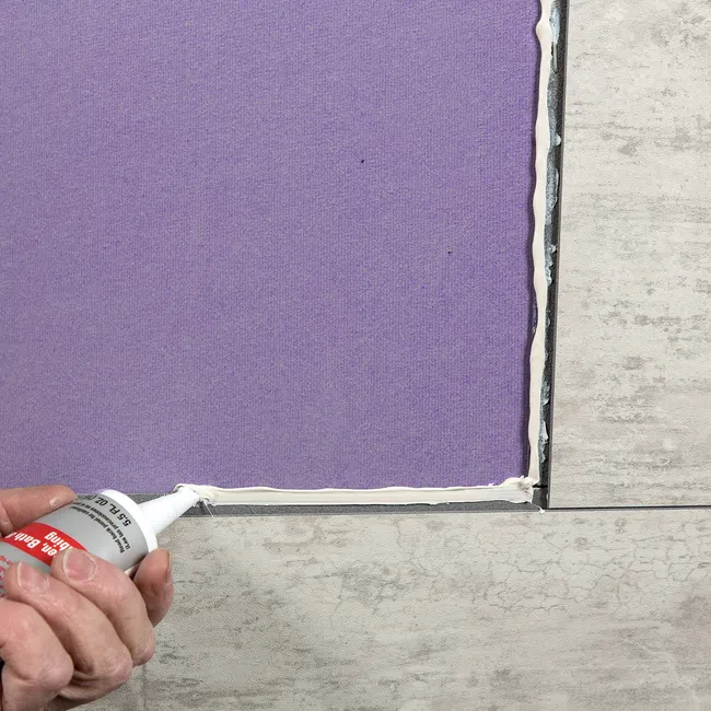 Hand applying white adhesive or sealant along the edge of a purple tile on a tiled floor corner.