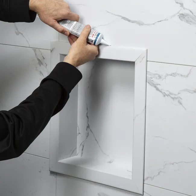 Person applying white sealant around a recessed shower niche framed with marble-effect tiles on a bathroom wall.