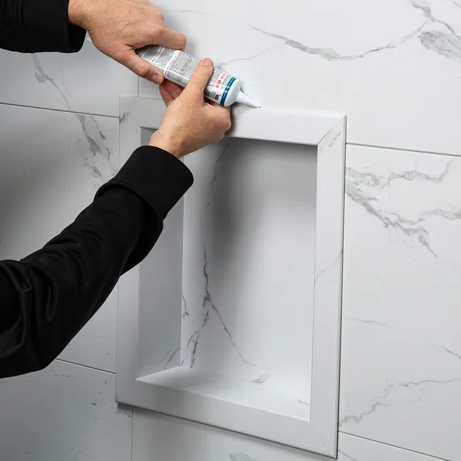 Person applying sealant to corner of recessed marble tiled shower niche on wall installation.