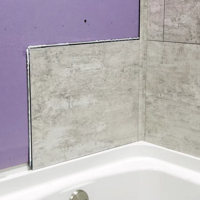Partially installed gray textured wall panels above a white bathtub with purple drywall visible behind.