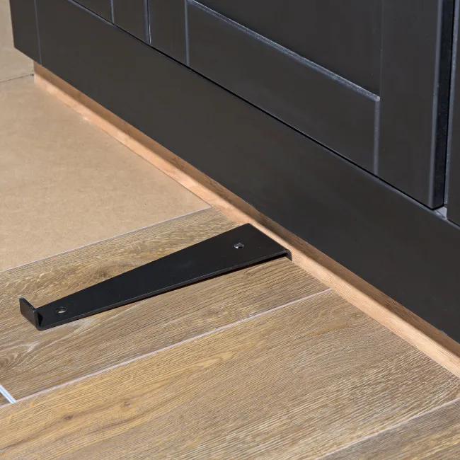 Black metal bracket on wood-look tile floor next to dark kitchen cabinetry baseboard.