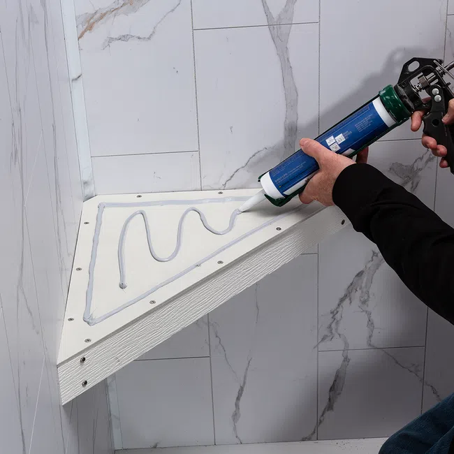 Person applying sealant with a caulking gun to a triangular shelf installed on a marble-patterned tiled wall.