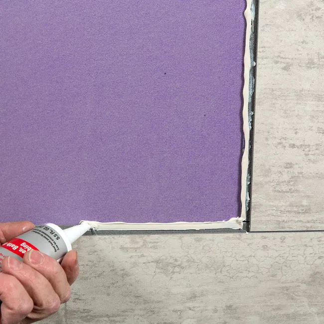 Hand applying white adhesive sealant around edge of purple tile on beige floor during installation