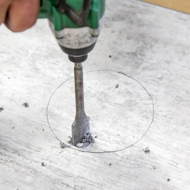 Electric drill with a flat bit creating a hole in a marked circular area on a gray stone surface.