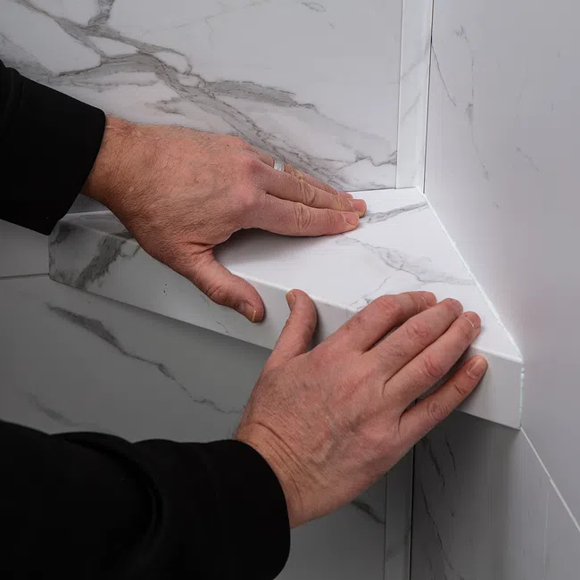 Hands installing white marble-patterned corner shelf on tiled wall with gray veining detail.