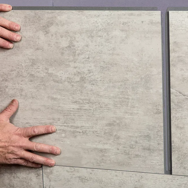 Two hands placing a large beige ceramic floor tile on a surface with grout lines visible.