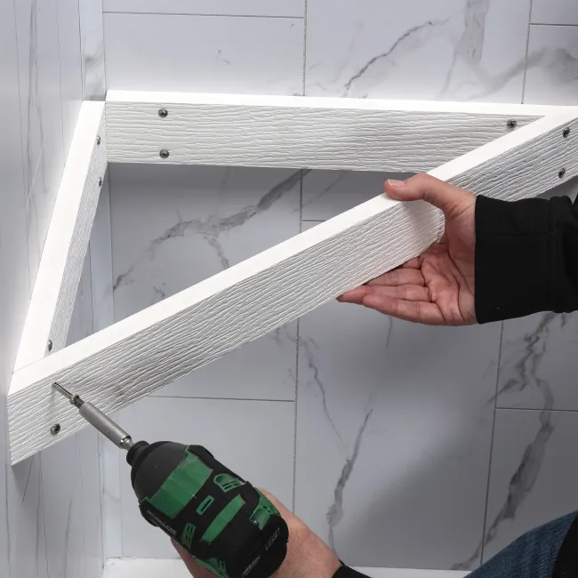 Person installing a white wooden shelf frame with a power drill on a marble wall background.