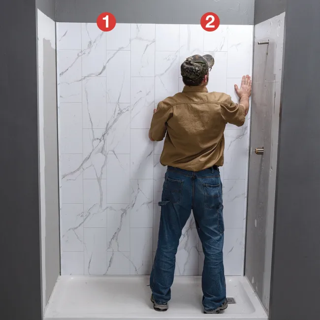 Man installing large white marble-effect tiles on shower wall in a bathroom under construction.