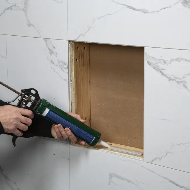 Applying white sealant to the edge of a cutout in marble wall tiles during home renovation.
