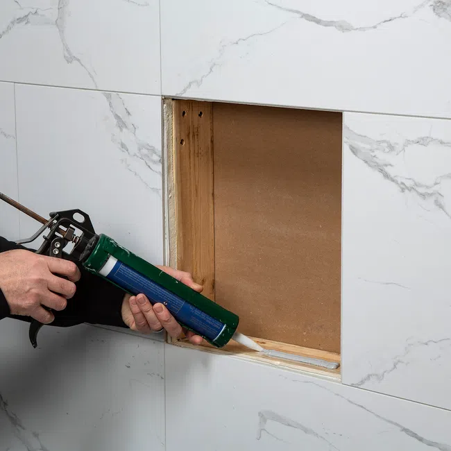 Person applying white sealant with a caulking gun to a wooden frame opening in marble tiled wall