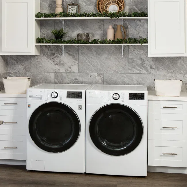 Modern laundry room with white front-load washer and dryer, gray tiled wall, white cabinets, and decorative shelves.
