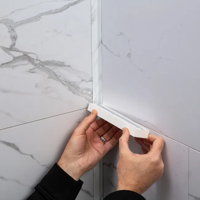 Hands installing a white corner tile trim piece on a marble-patterned tiled wall corner.