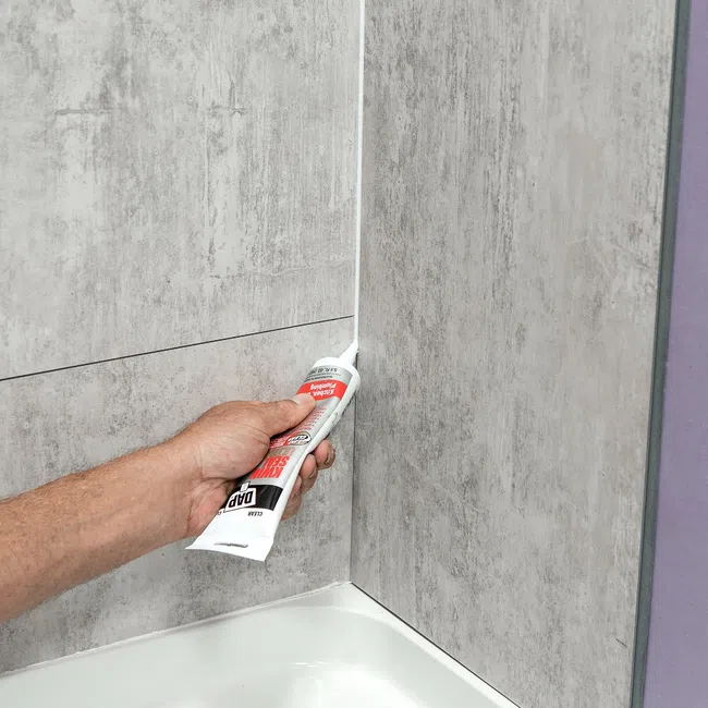 Hand applying white waterproof sealant caulk to bathtub corner between tiled walls.