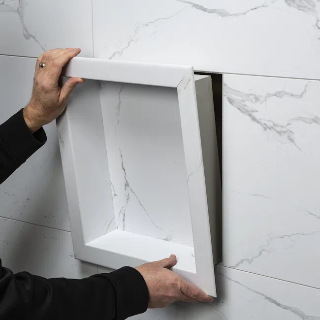 Hands installing a white marble recessed shower niche in a matching marble tiled wall.