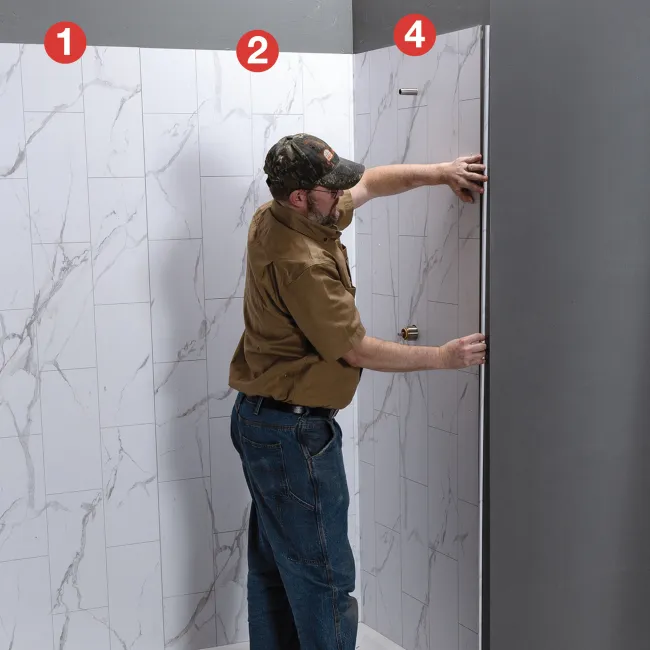 Man installing a glass shower door in a modern bathroom with white marble tiled walls and gray ceiling.