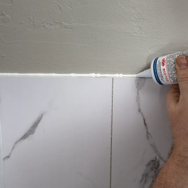 Hand applying white caulk along the edge where marble tiles meet a painted wall for sealing.