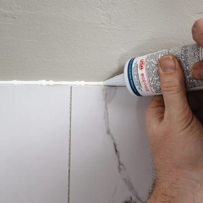 Hand applying white caulk between marble tiles and wall for sealing and finishing touch in home improvement.