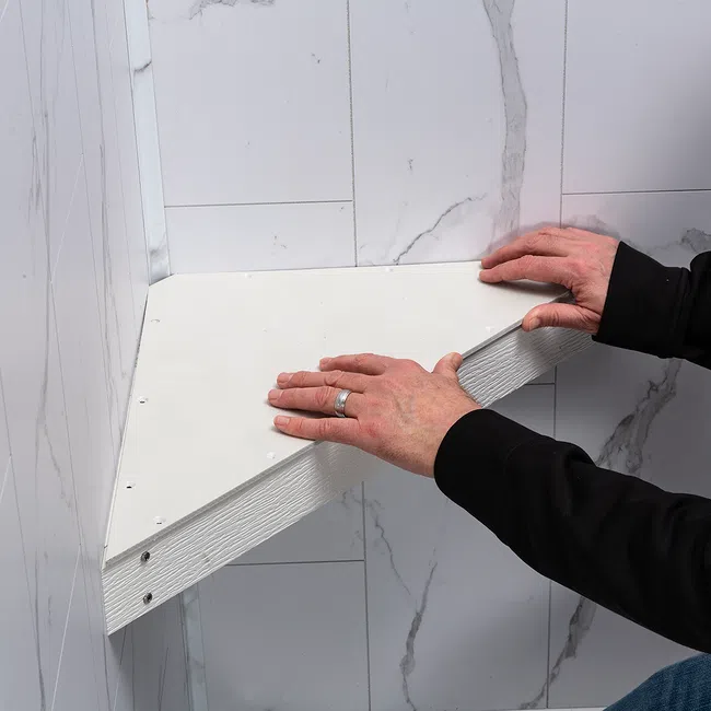Hands installing a white wooden corner shelf against a wall with white marble-patterned tiles.