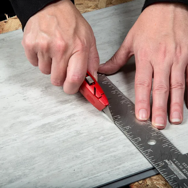 Person cutting a tile precisely using a red utility knife and metal square ruler on a work surface.