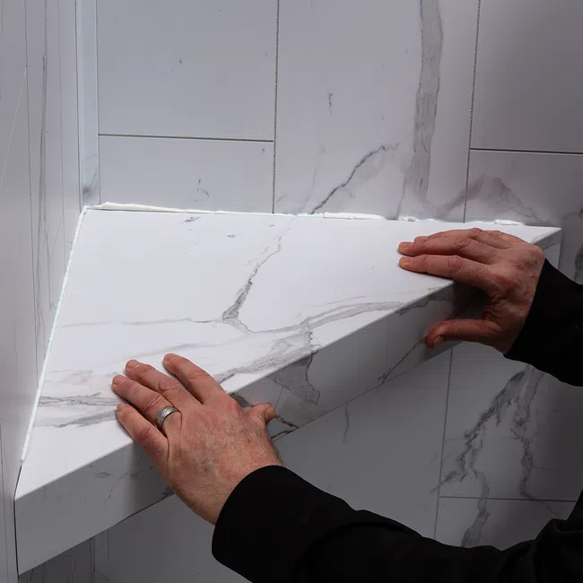 Hands installing a white marble corner shelf with gray veining on a tiled wall.