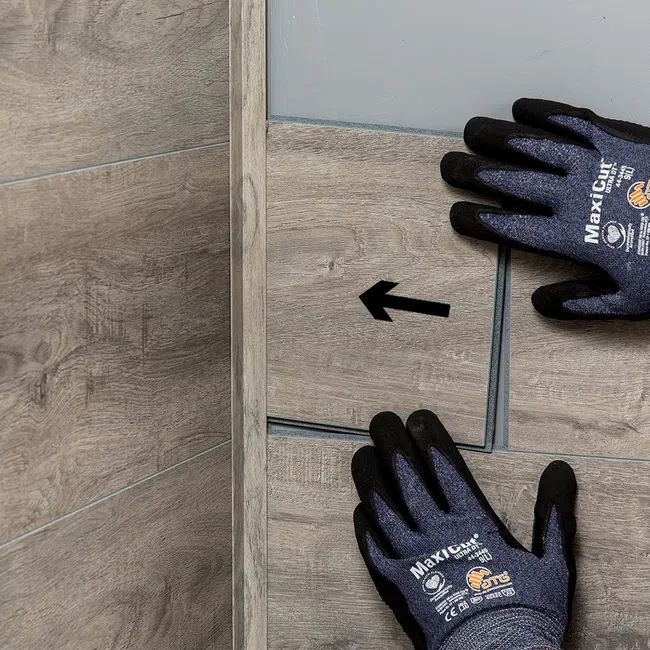 Hands with protective gloves installing wood-look floor tiles with directional arrows in a corner.