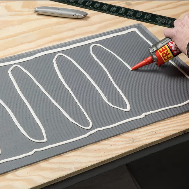 Applying white adhesive in zigzag pattern on a gray board resting on wooden surface with tools nearby