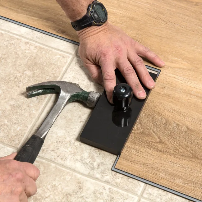 Hands using a tapping block and hammer to install wood laminate flooring over tile.