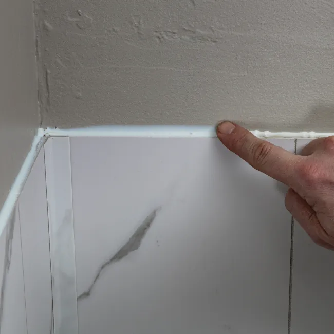 Hand applies white caulk along the edge where marble tiles meet a painted wall corner for sealing.